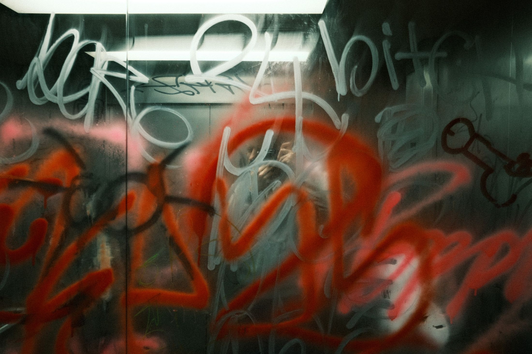 The mirrored wall of a lift, fogged and covered edge to edge in silver and red graffiti; the photographer's reflection is a ghost at its centre.