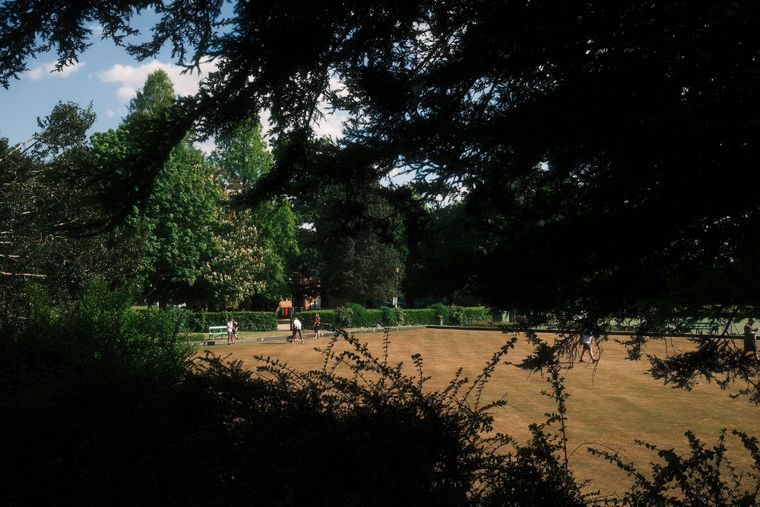 A lawn-bowls green seen from deep shade through overhanging branches; distant figures in whites play in the sun.