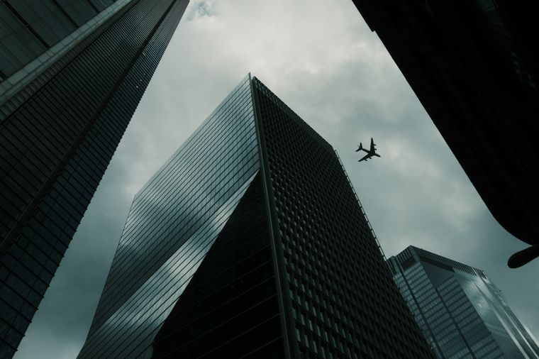 Glass towers seen from directly below, near-black against a bruised sky; a lone airliner crosses the gap between them.