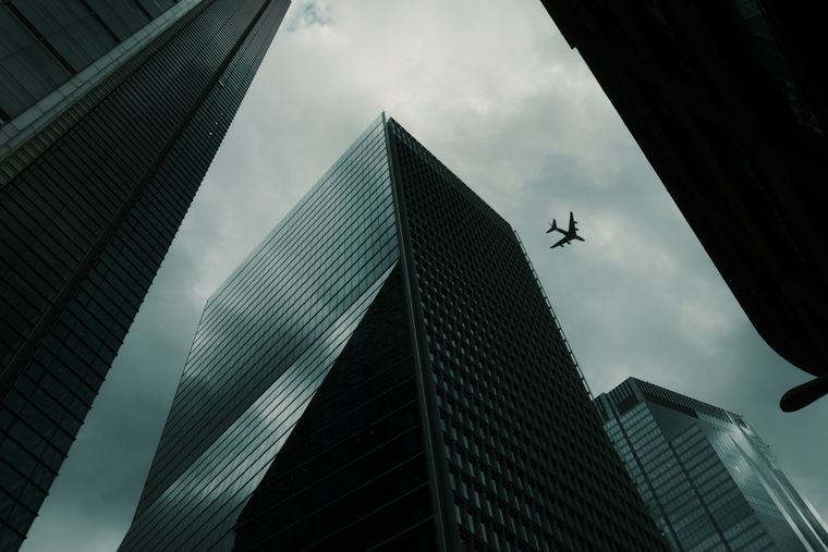 Glass towers seen from directly below, near-black against a bruised sky; a lone airliner crosses the gap between them.