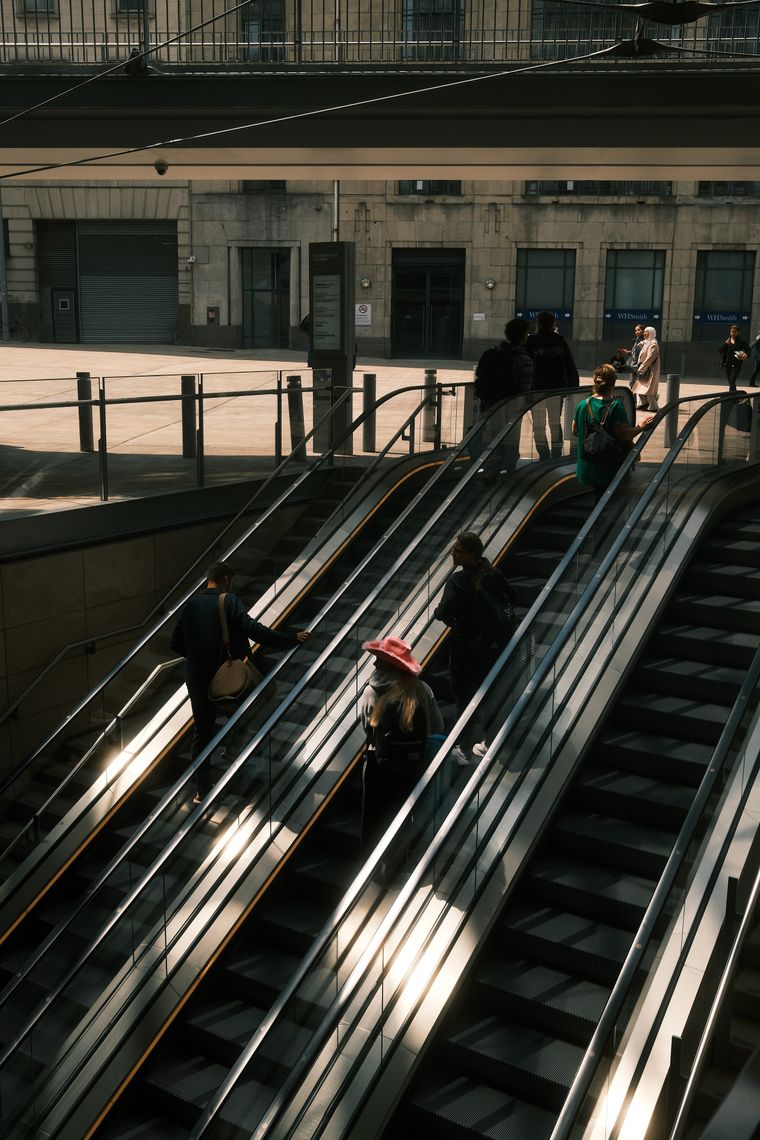 Long escalators descend into a station; a figure in a pink hat rides down through a blade of sunlight, stone facades above.